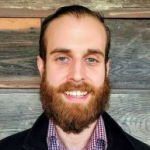 close up of a white man with a beard and mustache in a plaid shirt and a black blazer